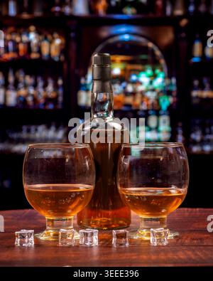 Deux verres à whisky arrondis, une boisson ambre, des glaçons et une bouteille sur un comptoir de bar en bois avec des étagères colorées de pub irlandais dans la douce mise au point. Banque D'Images