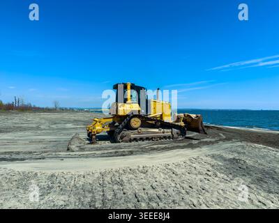 Un bulldozer jaune travaille sur une zone sablonneuse à côté du lac. Le soleil brille et l'eau est calme. Les gens peuvent être à proximité appréciant le temps. Banque D'Images
