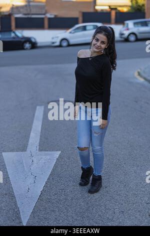 Jeune femme souriant décontractée tout en se tenant sur une route avec une flèche directionnelle Banque D'Images