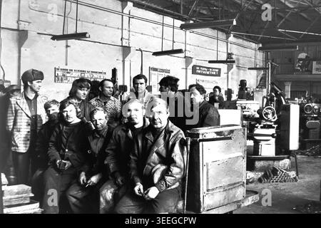 Slaviansk, 1983. À l'intérieur d'un atelier de l'armature-Insulator Factory (AIZ). Un groupe d'ouvriers d'usine masculins pose pour un portrait, assis et debout ensemble parmi les équipements industriels lourds. Cette photographie vintage en noir et blanc capture l'atmosphère authentique de l'époque, reflétant les activités quotidiennes et la culture industrielle de la région pendant la période soviétique. Il sert de document historique précieux de la communauté locale, préservant la mémoire visuelle des gens et de leur vie quotidienne, mettant en valeur un Donbass paisible. Banque D'Images