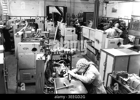 Slaviansk, 1983. À l'intérieur d'un atelier très fréquenté à l'usine d'armature-isolateur (AIZ). Une femme portant un foulard se concentre sur son travail sur un tour au premier plan, tandis que d'autres employés utilisent diverses machines en arrière-plan. Cette photographie vintage en noir et blanc capture l'atmosphère authentique de l'époque, reflétant les activités quotidiennes et la culture industrielle de la région pendant la période soviétique. Il sert de document historique précieux de la communauté locale, préservant la mémoire visuelle du peuple, mettant en valeur un Donbass pacifique. Banque D'Images