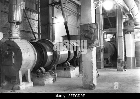 Slavyansk, 1983. Territoire de l'usine d'armature-isolant (AIZ). Photographie d'archives en noir et blanc capturant une scène historique fascinante. Les tambours cylindriques massifs en acier tournent régulièrement à l'intérieur de l'atelier d'usine expansif. Ces broyeurs à boulets mécaniques lourds ou sécheurs industriels sont connectés à des tuyaux épais et des conduits de ventilation, effectuant le traitement des matières premières essentielles pour la production de céramique. Cette image détaillée préserve un véritable fragment de l'histoire industrielle soviétique, mettant en valeur la vie avant la guerre et le Donbass pacifique. Banque D'Images