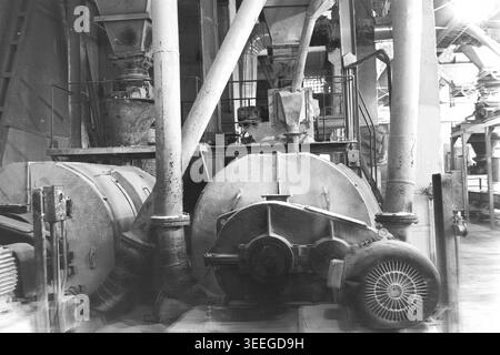 Slavyansk, 1983. Territoire de l'usine d'armature-isolant (AIZ). Photographie d'archives en noir et blanc capturant une scène historique fascinante. Les tambours cylindriques massifs en acier tournent régulièrement à l'intérieur de l'atelier d'usine expansif. Ces broyeurs à boulets mécaniques lourds ou sécheurs industriels sont connectés à des tuyaux épais et des conduits de ventilation, effectuant le traitement des matières premières essentielles pour la production de céramique. Cette image détaillée préserve un véritable fragment de l'histoire industrielle soviétique, mettant en valeur la vie avant la guerre et le Donbass pacifique. Banque D'Images
