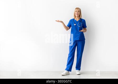 Médecin féminin mature dans les gommages bleus présentant l'espace de copie sur fond blanc Banque D'Images