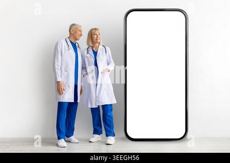 Deux médecins matures en blouses de laboratoire debout à côté de l'écran blanc géant de smartphone Banque D'Images