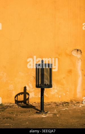 Une poubelle de rue en métal noir jette une ombre nette sur un mur orange ensoleillé et altéré, produisant une composition urbaine minimaliste avec une texture riche, qu Banque D'Images