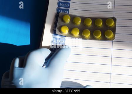 Une main dans un gant médical bleu pointant vers un calendrier de médicaments dans un planificateur quotidien à côté de pilules jaune vif à la lumière du soleil. Banque D'Images