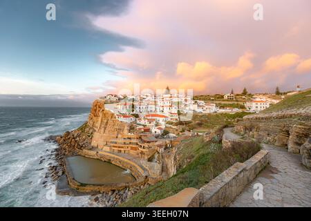 Village côtier cinématographique perché sur des falaises au coucher du soleil. Les maisons blanches brillent chaleureusement contre le ciel pastel. Les vagues s'écrasent près de chemins de pierre et de murs anciens. Banque D'Images