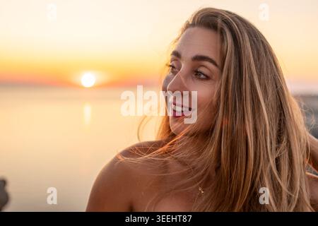 Femme coucher de soleil plage souriante jeune dame regarde loin à la mer pendant l'heure dorée en vacances appréciant le voyage et les moments de la nature paisible Banque D'Images