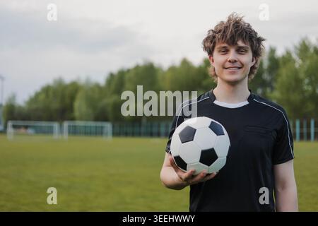 Jeune homme souriant en uniforme de football noir tenant un ballon de football sur un terrain. Copier l'espace. Banque D'Images
