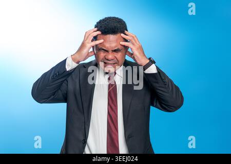 Portrait d'un homme indonésien stressé dans un costume tenant sa tête dans la douleur. Studio tourné sur fond bleu représentant le stress de l'entreprise ou la dette. Banque D'Images