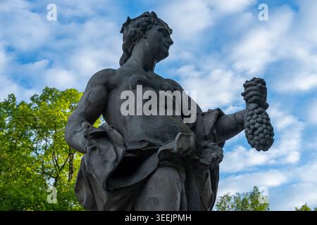 Statue classique en pierre d'une figure mythologique tenant des raisins dans un cadre de parc verdoyant. Banque D'Images