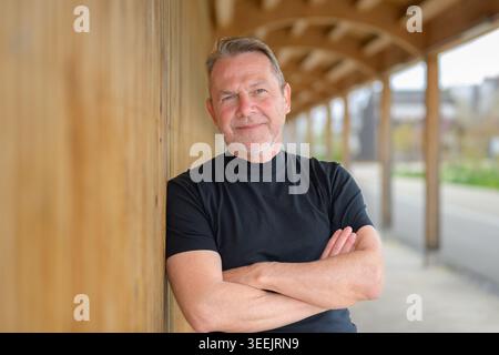 Portrait d'un homme d'affaires mûr détendu penché avec désinvolture contre un mur en bois dans l'architecture moderne. Style de vie professionnel, confiance et authenticité Banque D'Images