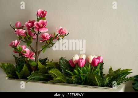 Magnifique magnolia rose artificiel et tulipes roses avec des fougères vertes disposées dans une jardinière contre un mur beige Uni. Banque D'Images