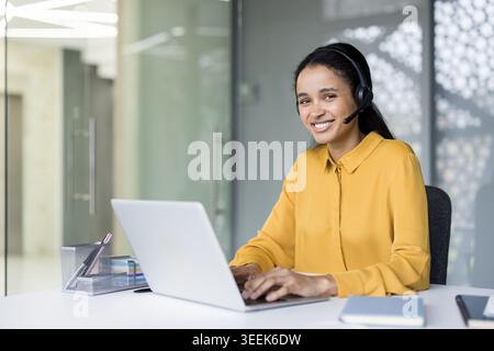 Jeune femme souriante offrant un service client amical et une assistance à distance depuis un bureau moderne, portant un casque noir tout en travaillant en toute confiance sur un ordinateur portable Banque D'Images