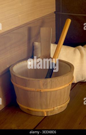 Un seau de sauna finlandais classique en bois avec une doublure en plastique et une louche assortie reposant sur un banc en bois. La scène comprend un oreiller de sauna moelleux. Banque D'Images