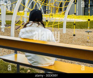 Jeune femme dans une veste blanche s'assoit tranquillement sur un banc, regardant vers une aire de jeux moderne avec un équipement d'escalade lumineux Banque D'Images