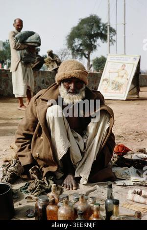 Une diapositive de voyage d'archives vintage en couleur du début des années 1970 représentant un homme indien âgé portant un manteau épais et un couvre-chef, assis à l'extérieur sur le sol dans une zone de marché de New Delhi. Banque D'Images
