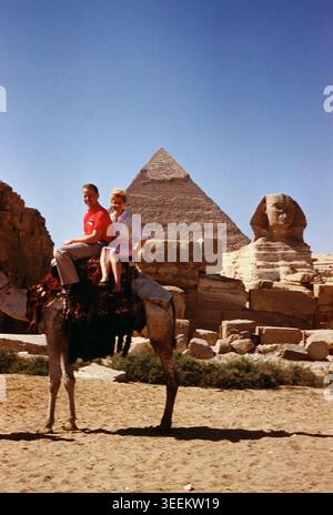 Touristes montant un chameau guidé par un guide égyptien local, positionné sur une piste en terre devant les pyramides de Gizeh sous un soleil désertique éclatant et éblouissant. Banque D'Images