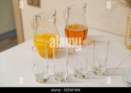 Deux pichets en verre avec des boissons à base d'orange et de thé glacé se tiennent derrière de grands verres vides sur une nappe neutre. Banque D'Images