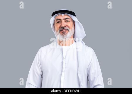 Homme arabe mûr posant sur un fond gris, exprimant la confiance et une disposition amicale, portant des vêtements traditionnels du moyen-Orient includin Banque D'Images