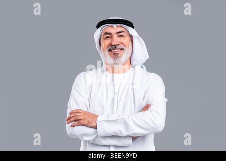 Homme arabe senior portant la kandora et le ghutra émiratis traditionnels, souriant avec confiance avec les bras croisés, représentant la sagesse, la tradition et le succès Banque D'Images