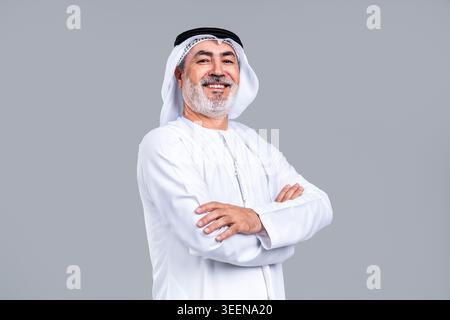 Homme arabe senior en kandora et ghutra émirati traditionnels avec agal, souriant avec confiance avec les bras croisés dans un portrait de studio contre un gris neutre Banque D'Images