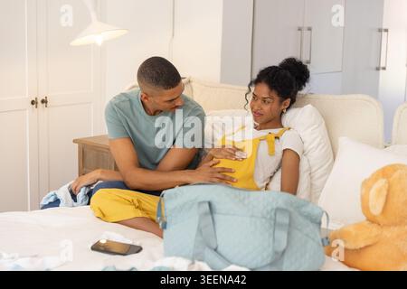 Couple afro-américain assis sur le lit, femme portant une salopette jaune, homme touchant le ventre, sac bébé Banque D'Images