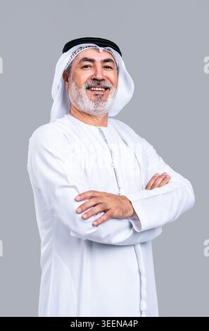 Homme arabe mature senior portant la kandora émiratie traditionnelle, le ghutra et l'agal souriant avec confiance et les bras croisés, dépeignant la sagesse et le cul fier Banque D'Images
