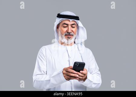 Homme arabe senior dans la traditionnelle kandora émirati et ghutra souriant tout en s'engageant avec un smartphone, représentant le style de vie numérique moderne, communicati Banque D'Images