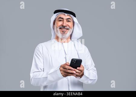 Homme arabe senior portant la kandora et le ghutra émiratis traditionnels, souriant avec confiance tout en tenant et en interagissant avec un smartphone, représentant le mode Banque D'Images