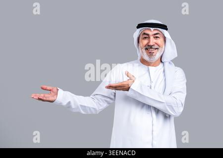 Homme arabe mature senior en kandora, ghutra et agal émiratis traditionnels, souriant largement et faisant des gestes avec les mains ouvertes, indiquant un accueil et un pré Banque D'Images