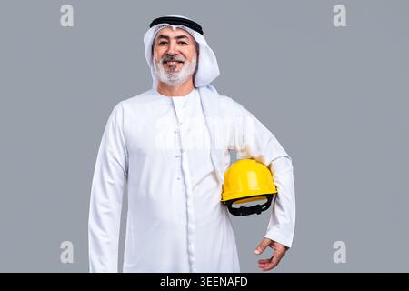 Homme émirati senior portant des kandora et ghutra traditionnels, souriant avec confiance tout en tenant un casque jaune, représentant l'expérience et la profession Banque D'Images