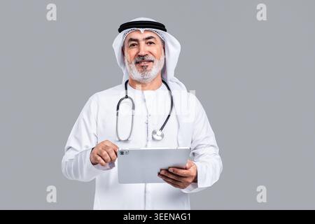 Médecin homme arabe senior portant des vêtements traditionnels du moyen-Orient et un stéthoscope, tenant en toute confiance une tablette numérique, représentant la technologie de la santé Banque D'Images