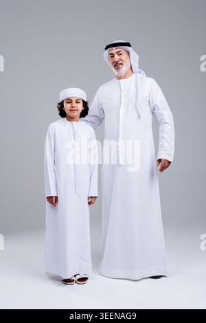 Grand-père émirati debout avec son petit-fils. Tous deux portent des kandora et ghutra traditionnels sur un fond gris clair. Symbolisant les liens familiaux. Trad Banque D'Images