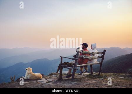 Deux personnes se reposant sur un banc en bois avec un chien regardant la vue sur la montagne au coucher du soleil Raghupur Fort, Sojha, Manali, Himachal Pradesh, Inde. Banque D'Images
