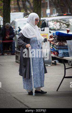 Une femme musulmane modestement habillée avec une canne et un hijab blanc fait des magasins sur un stand en plein air sur la 37e avenue à Jackson Heights, Queens à Little Bangaldesh Banque D'Images