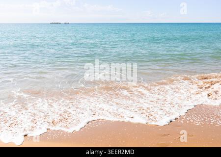 De douces vagues de l'océan se jettent sur une plage de sable, créant de la mousse le long du rivage. Le ciel bleu clair s'étend au-dessus, avec quelques rochers lointains visibles Banque D'Images