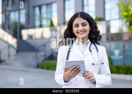 Jeune professionnel de la santé confiant portant une blouse blanche et un stéthoscope, utilisant une tablette tout en se tenant debout à l'extérieur d'un établissement de santé moderne, représentant l'innovation et le dévouement en médecine Banque D'Images