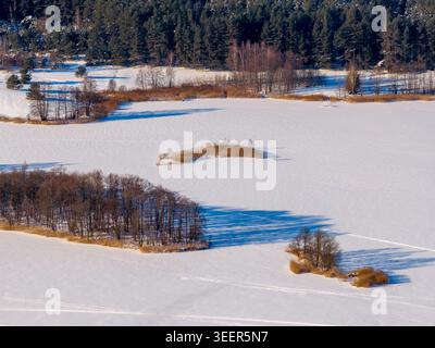 Vue aérienne d'un lac gelé avec de petites îles d'arbres et une forêt dense à feuilles persistantes sous la lumière du soleil d'hiver en Lituanie. Banque D'Images