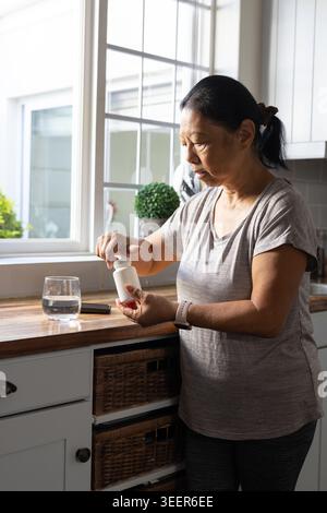 Femme asiatique senior debout au comptoir en bois ouvrant la bouteille de pilule blanche par verre d'eau Banque D'Images