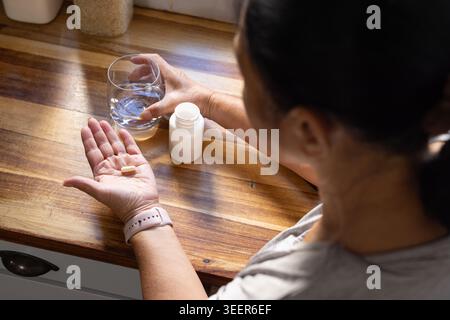 Femme asiatique senior montrant la pilule dans la paume tout en tenant le verre et la bouteille de pilule à côté du comptoir de cuisine Banque D'Images