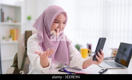 Femme d'affaires musulmane asiatique dans le hijab ayant un appel vidéo sur smartphone tout en travaillant au bureau à domicile avec ordinateur portable et Banque D'Images
