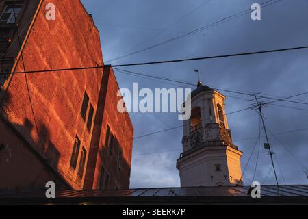 La tour de l'horloge historique de Vyborg s'élève à côté d'un bâtiment en briques rouges tandis que la lumière chaude du soleil à l'heure dorée illumine la façade, contrastant avec les nuages d'humeur changeante Banque D'Images