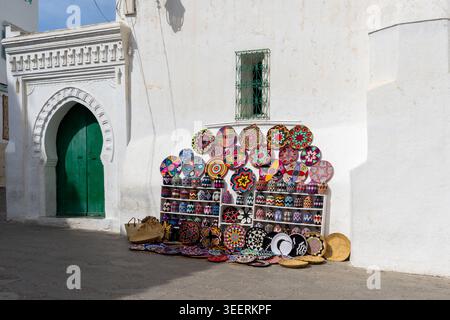 Souvenirs colorés affichés contre un mur blanc avec une porte traditionnelle peinte en vert avec un cadre voûté dans la vieille ville d'Asilah, au Maroc. Banque D'Images