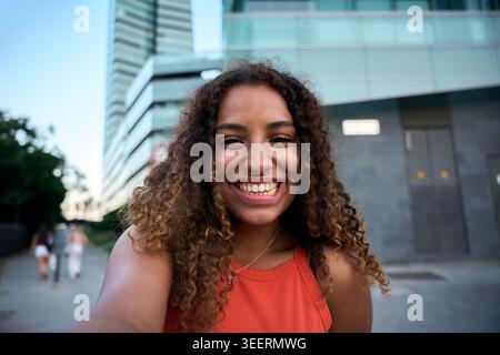 Femme noire souriante avec des cheveux bouclés dans un cadre urbain moderne par une journée ensoleillée Banque D'Images