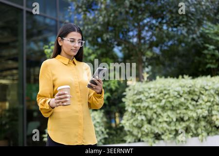 Jeune femme professionnelle portant des lunettes et une chemise jaune, tenant une tasse à café et envoyant des SMS sur un smartphone, debout à l'extérieur dans un environnement urbain avec des immeubles de bureaux et de la verdure Banque D'Images