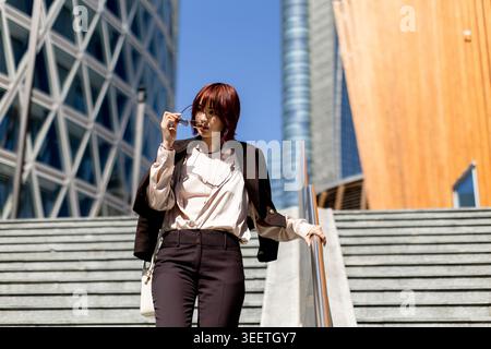 Une femme élégante descend en toute confiance les marches modernes, portant des lunettes de soleil, avec une architecture saisissante autour d'elle par une journée ensoleillée. Banque D'Images