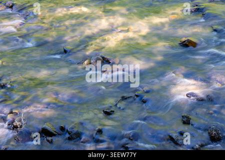 L'eau claire coule sur les pierres lisses. La lumière du soleil brille par de légères ondulations. Les ombres dansent à travers les rochers mouillés ci-dessous. Le rythme tranquille de la nature bouge avec Banque D'Images