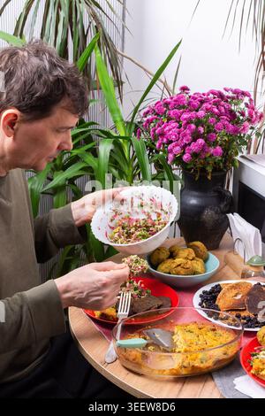 Un homme est assis à une table ronde et mange divers plats préparés à partir de recettes faites maison. Il est entouré de plantes et d'un bouquet de fleurs dans une cuisine ensoleillée pendant l'heure du petit déjeuner Banque D'Images